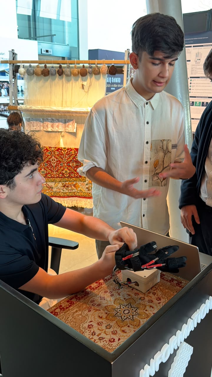 Close view of the ReHand Loom wearable prototype, a sensor glove wired to a small controller housing, being fitted onto a hand beside a carpet loom